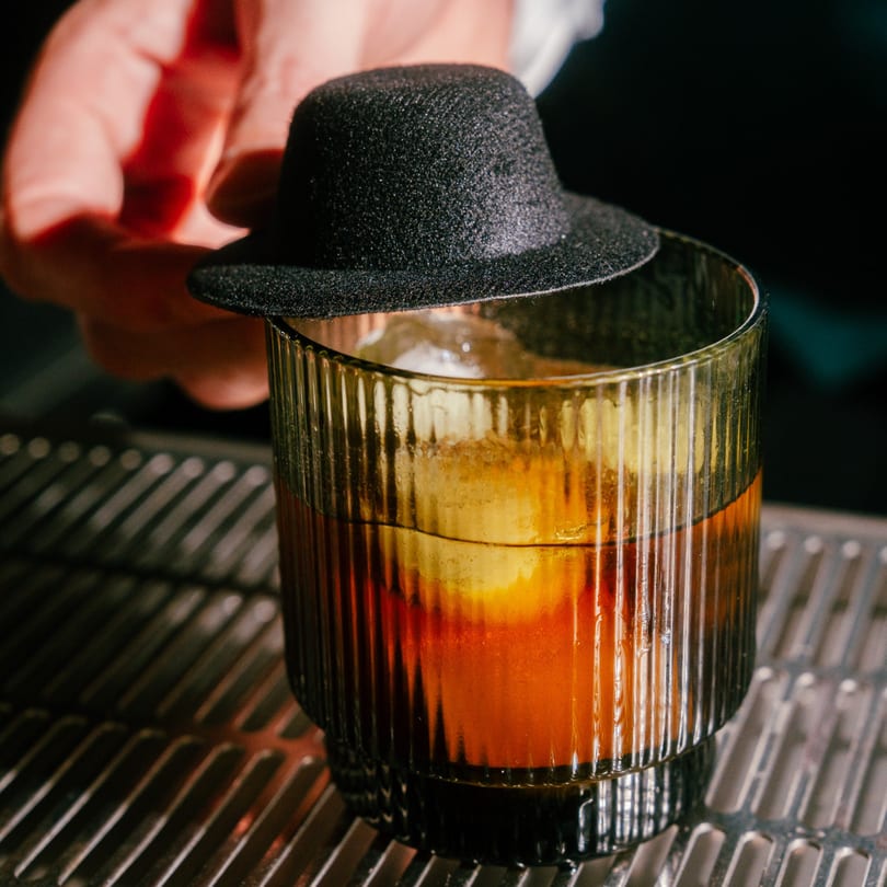 A hand places a miniature black fedora hat on top of a cocktail in a ribbed glass.
