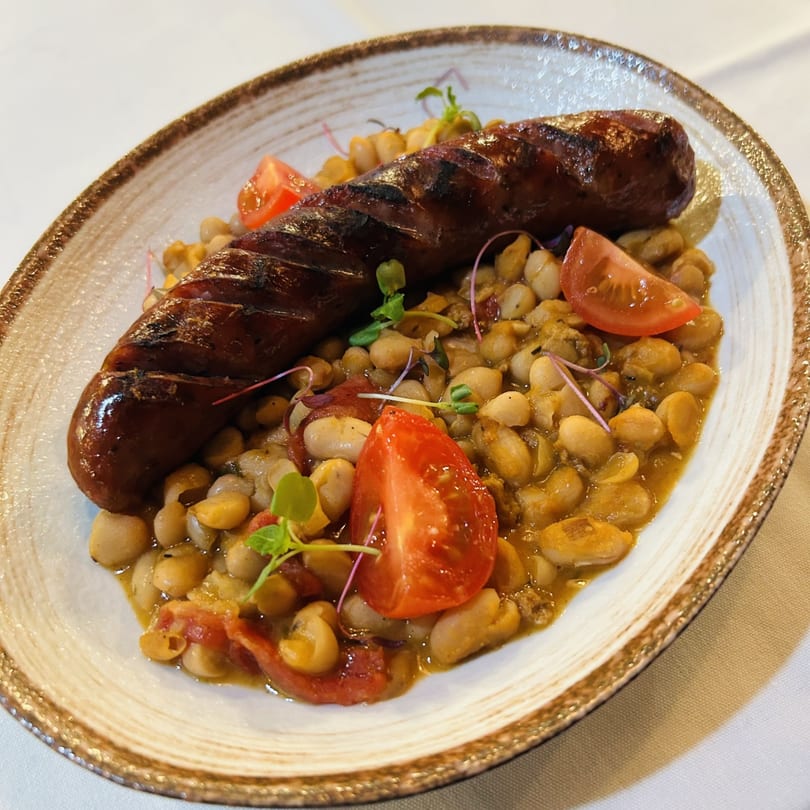 Grilled Italian sausage with white beans, cherry tomatoes, and microgreens on a rustic plate.